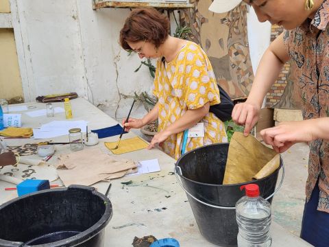 One woman soaking leather into a bucket of indigo dye solution and another using a brush to paint on designs to the leather.